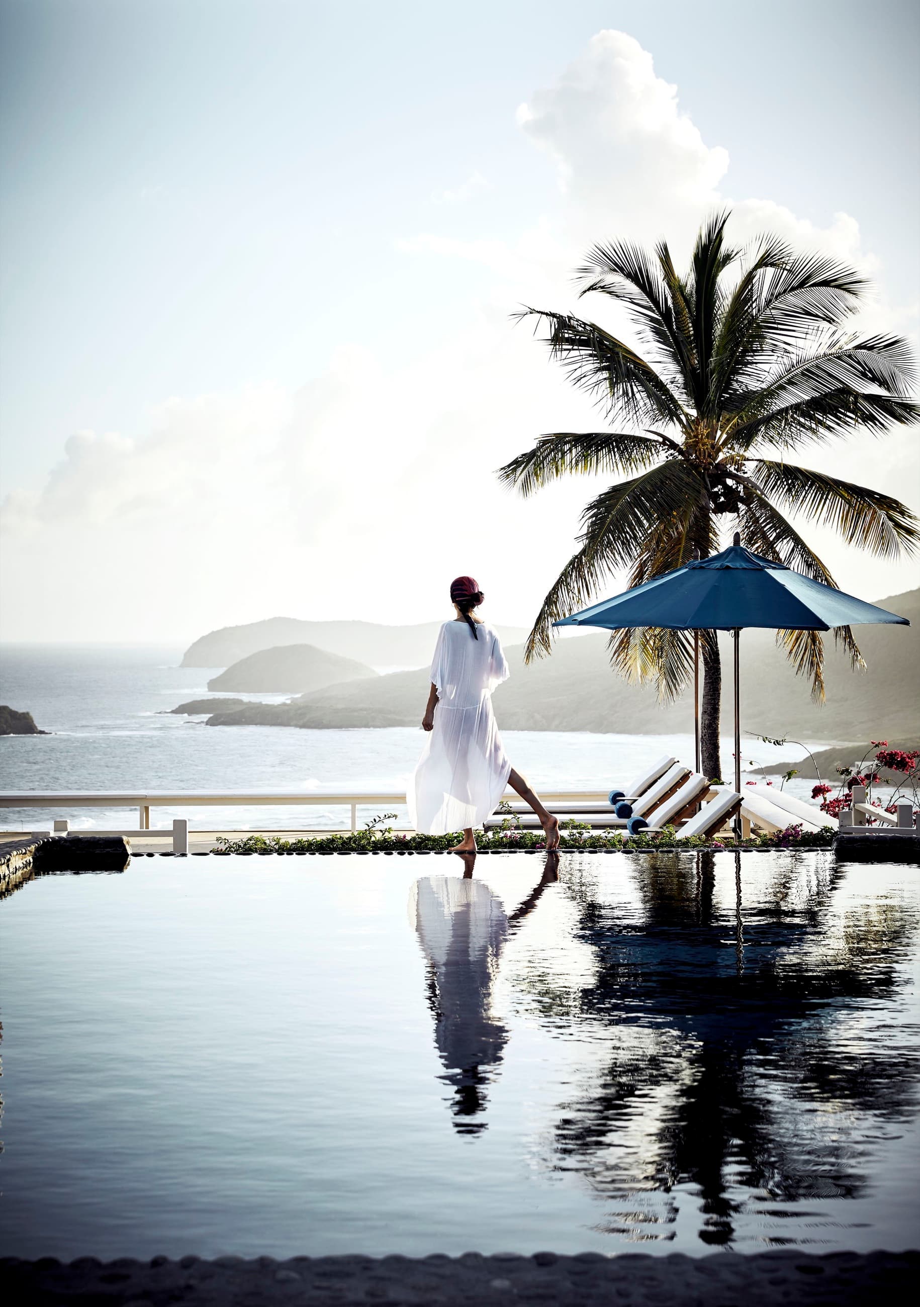 A woman in white walks the edge of an infinity pool beneath a palm